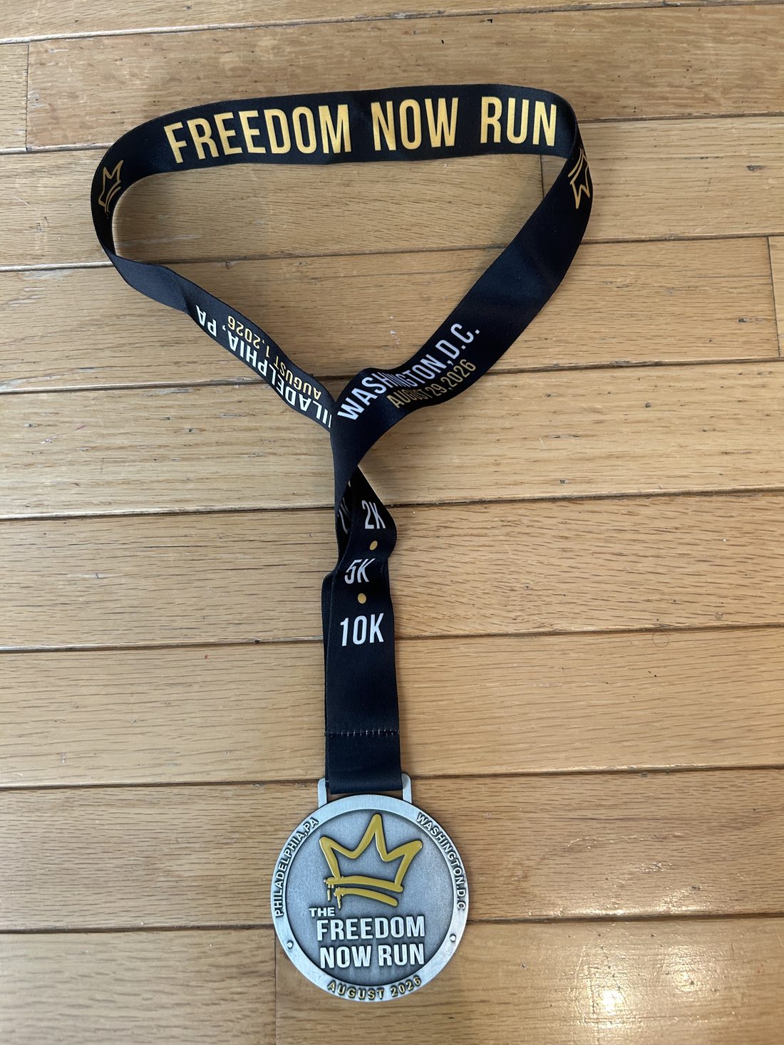 Front of the 2026 Freedom Now Run finisher medal, a silver disc with a gold crown logo reading The Freedom Now Run, August 2026, with Philadelphia PA and Washington DC stamped around the rim, on a black ribbon printed with 2K, 5K and 10K